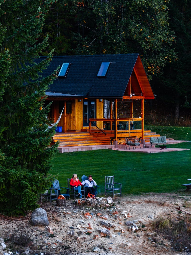 Wooden cabin with a lawn and Adirondack chairs above a rocky lakeshore in the forest at Kootenay Lakeview Retreats