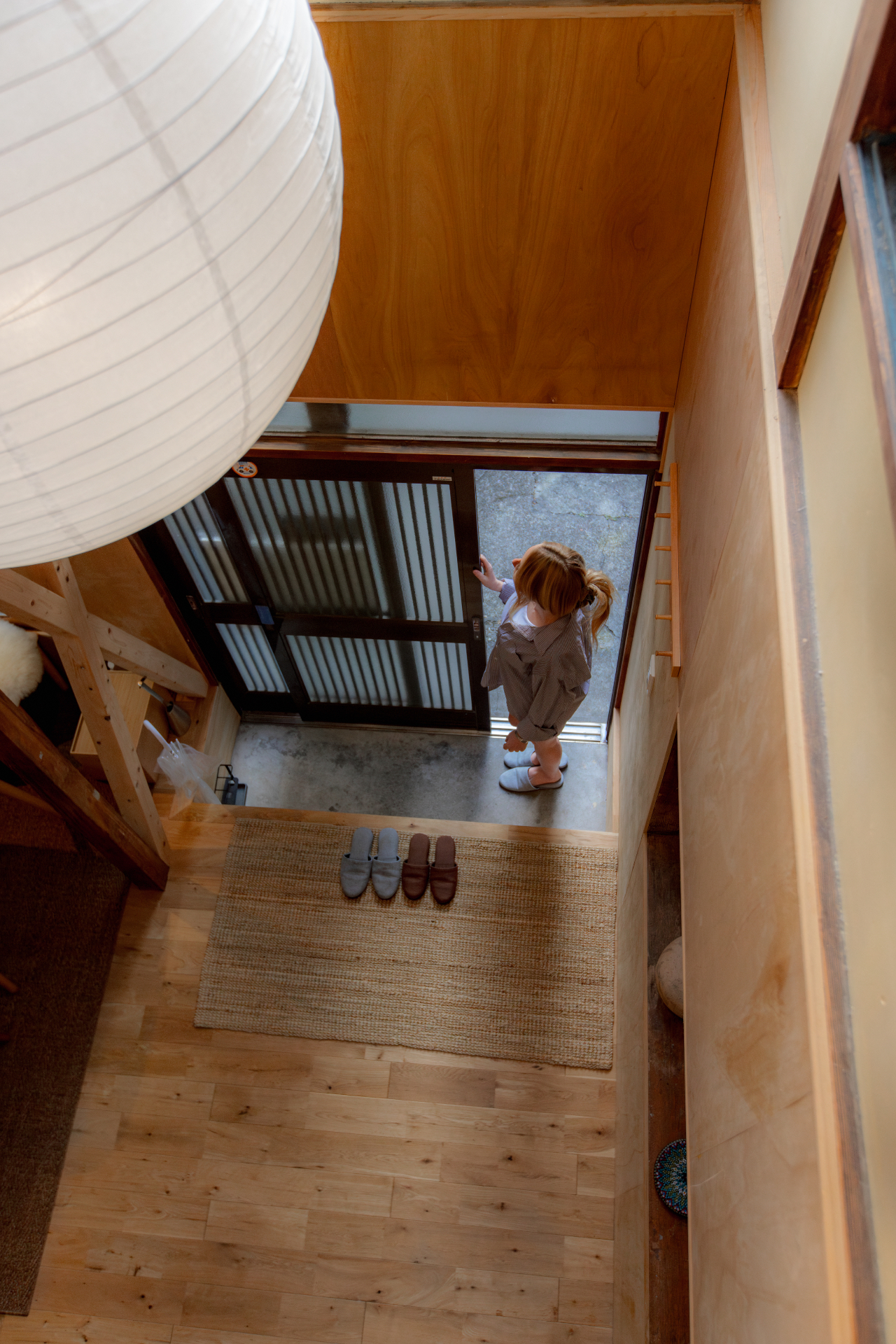 Overhead view of a traditional Japanese entryway with slippers on a mat, a large paper lantern, and a guest at the shoji front door