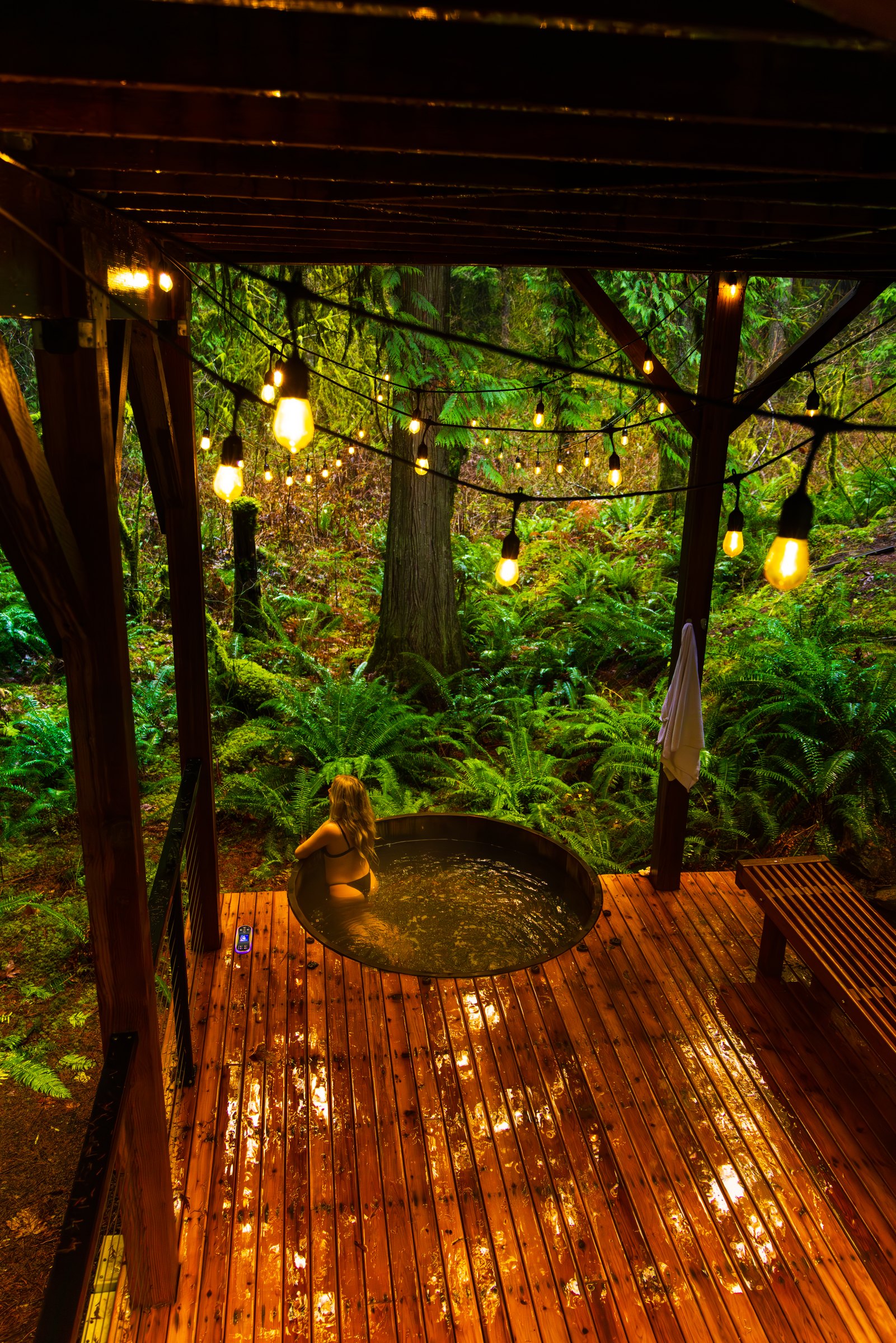 Guest in a round hot tub on a wet wooden deck under string lights, surrounded by ferns and tall trees at Mt Baker Hygge Haus
