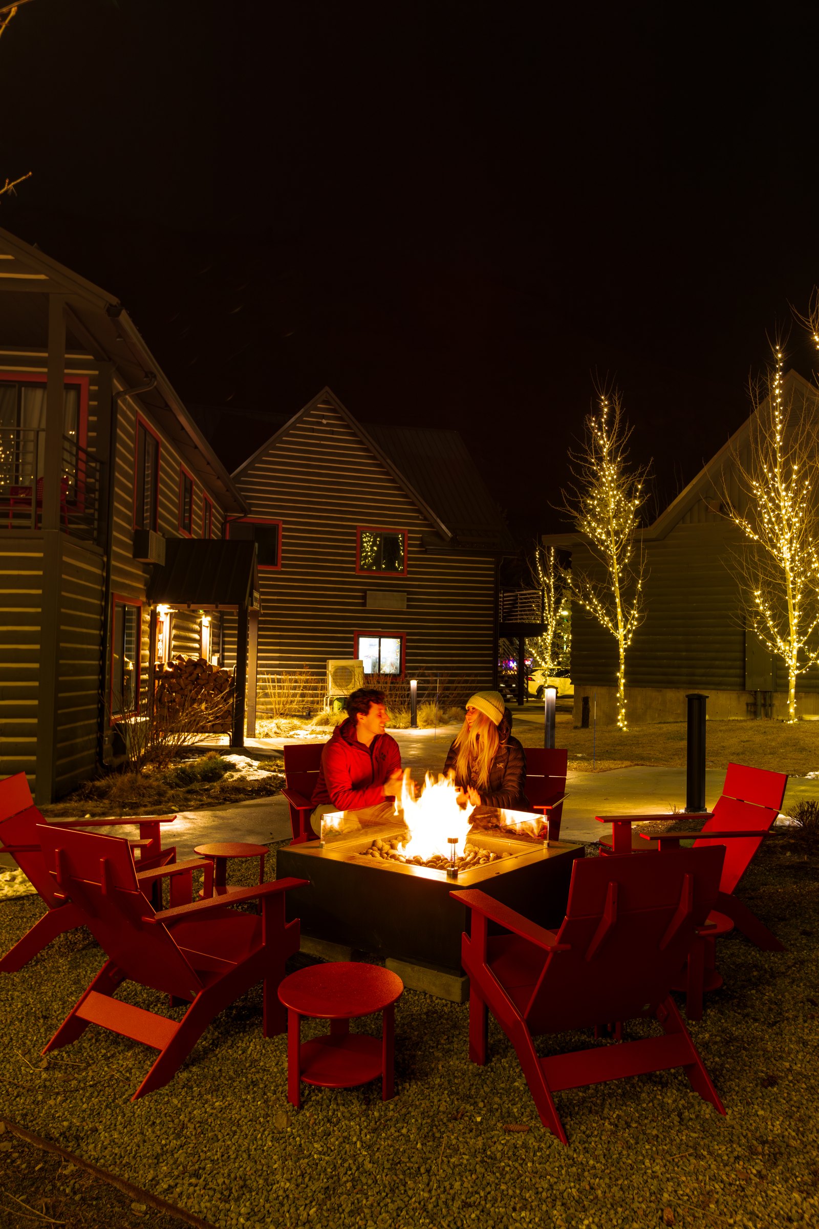 Two guests by the fire pit on red Adirondack chairs among string lit trees at Mountain Modern Motel