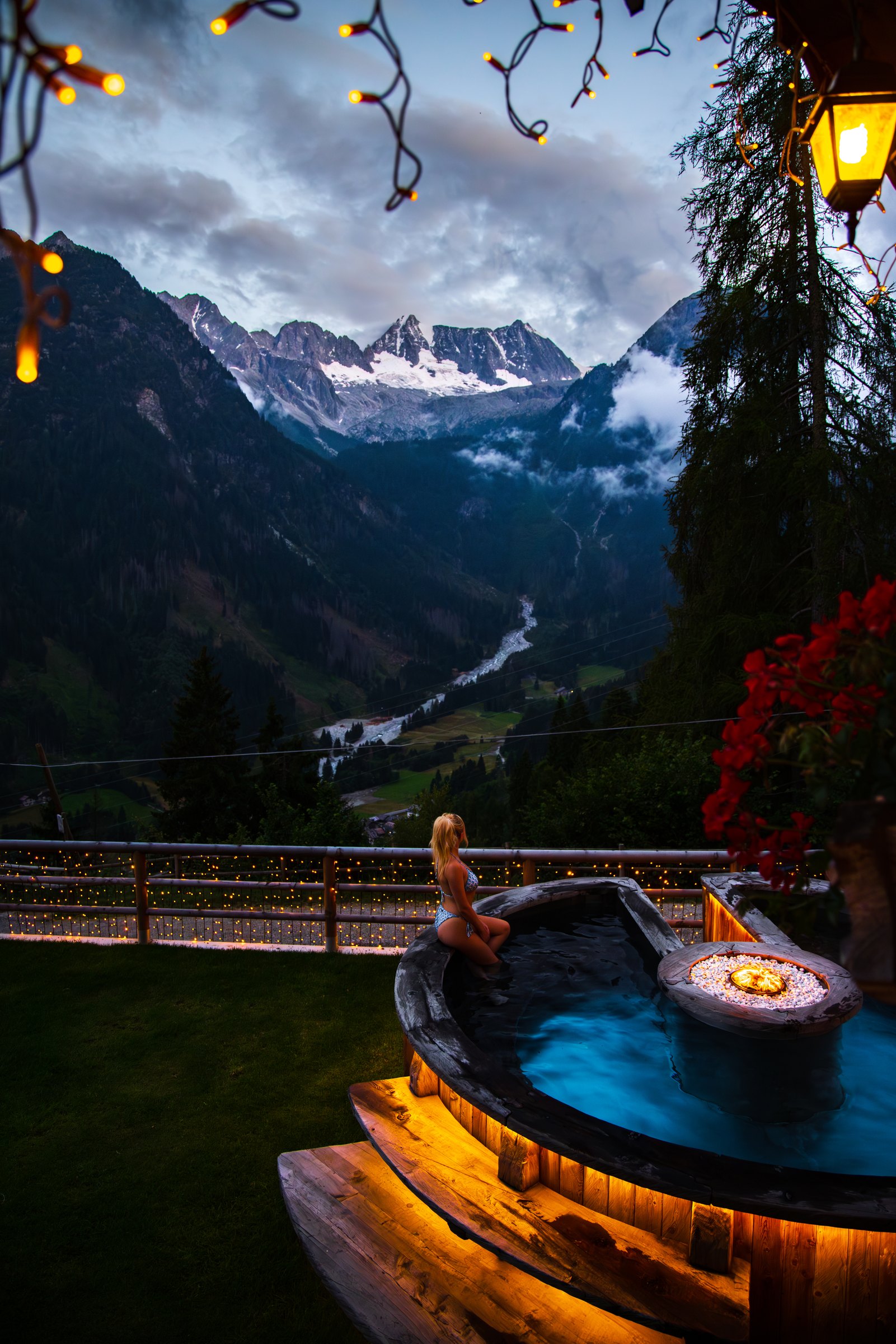 Wooden hot tub glowing under string lights with snowy peaks and a valley behind at Chalet Al Foss