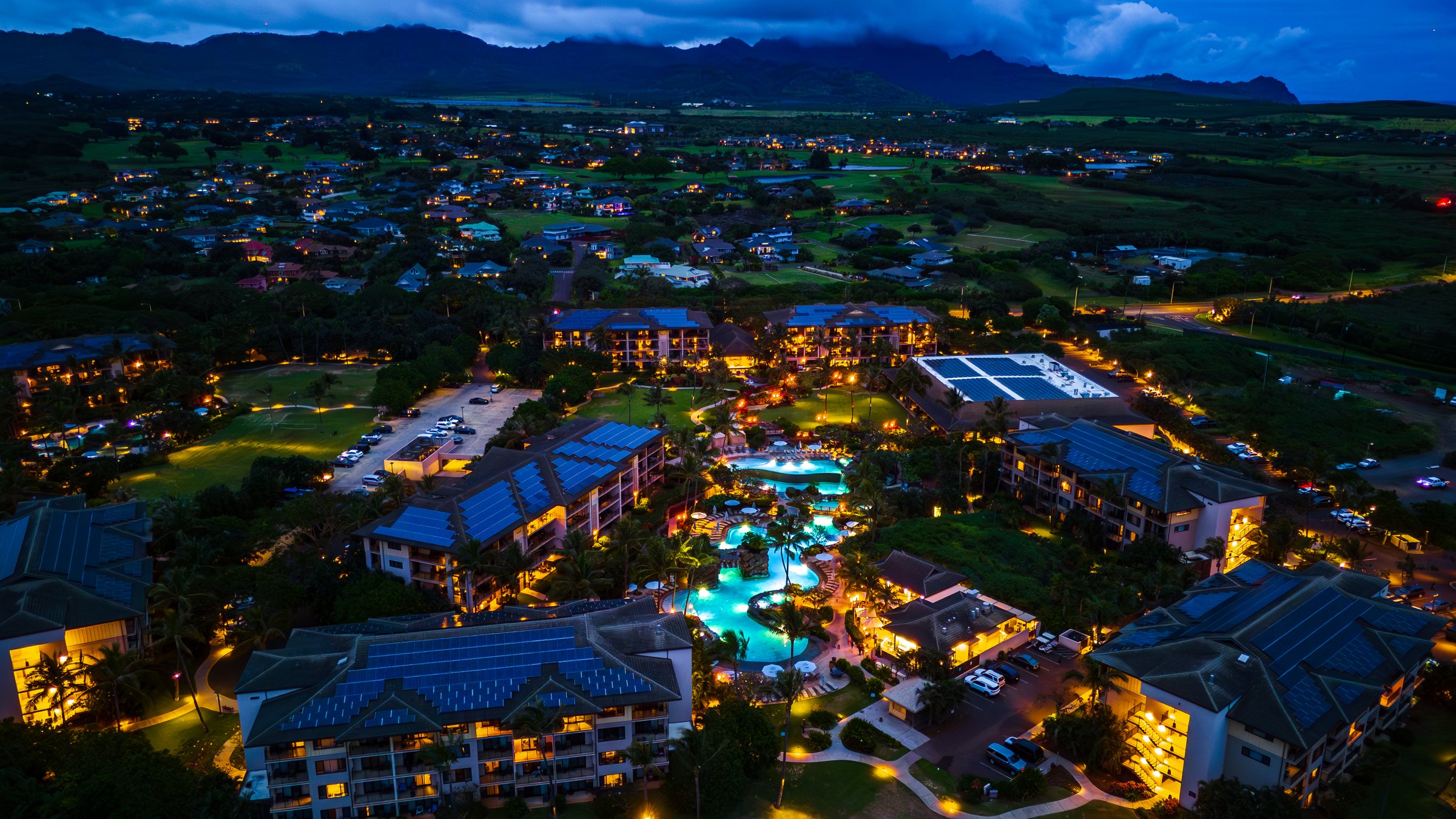 Aerial of Koloa Landing's glowing pools and buildings at blue hour with mountains behind