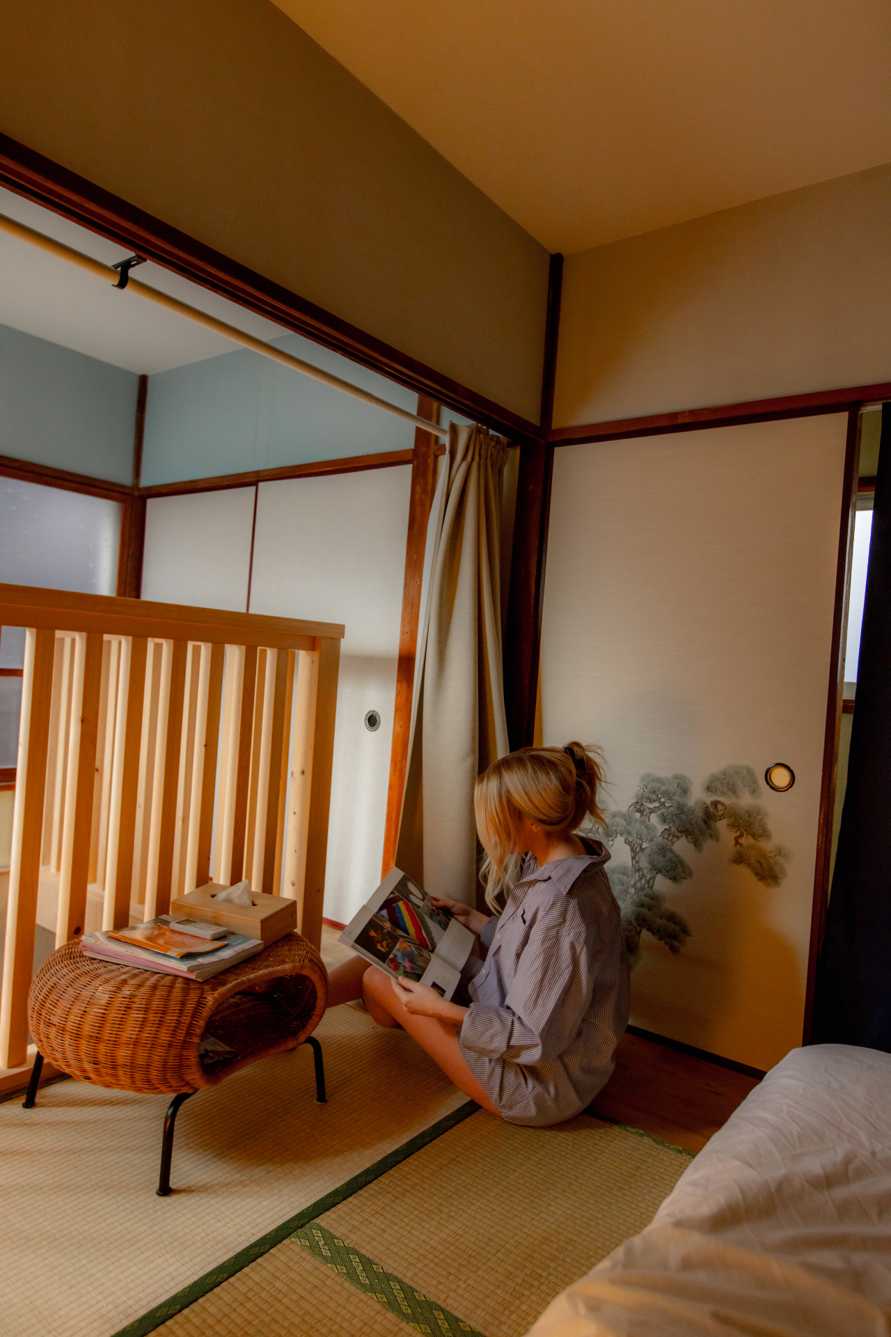 Guest reading a magazine on tatami beside a pine-painted shoji door and a woven stool in a traditional Japanese home