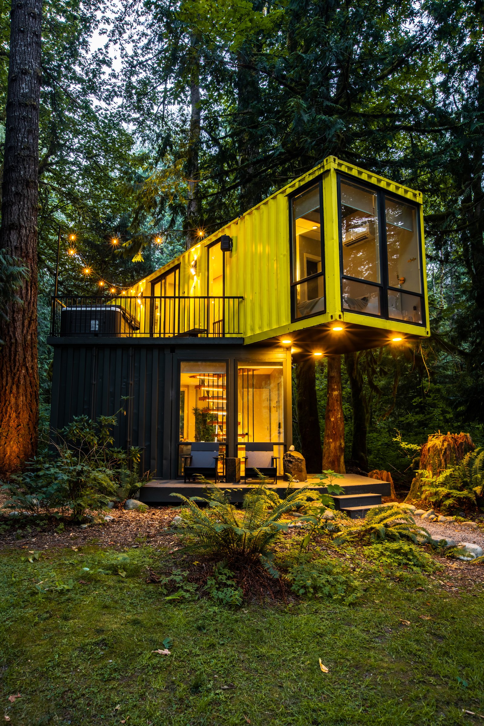 Yellow container cabin glowing at dusk among tall trees at Index Cabins