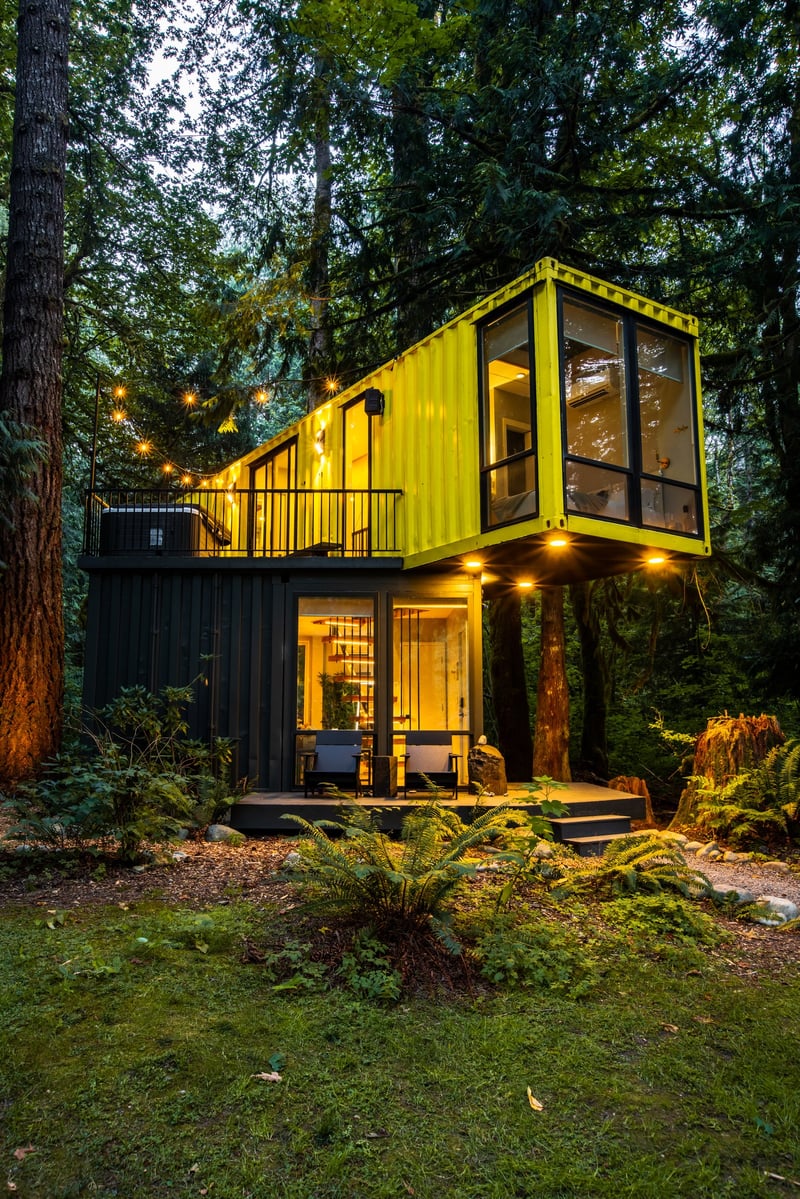 Yellow container cabin glowing at dusk among tall trees at Index Cabins