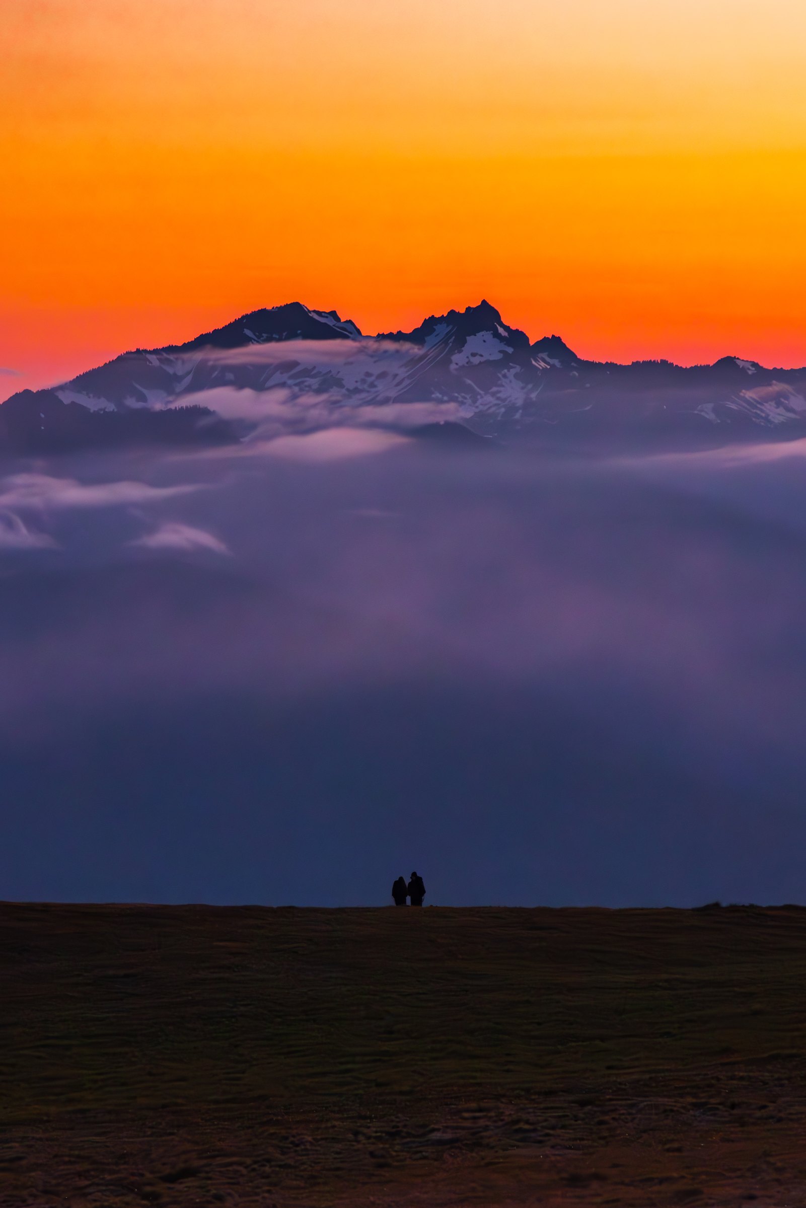 Two people silhouetted on Hurricane Ridge below snowy Olympic peaks and a fiery orange sky