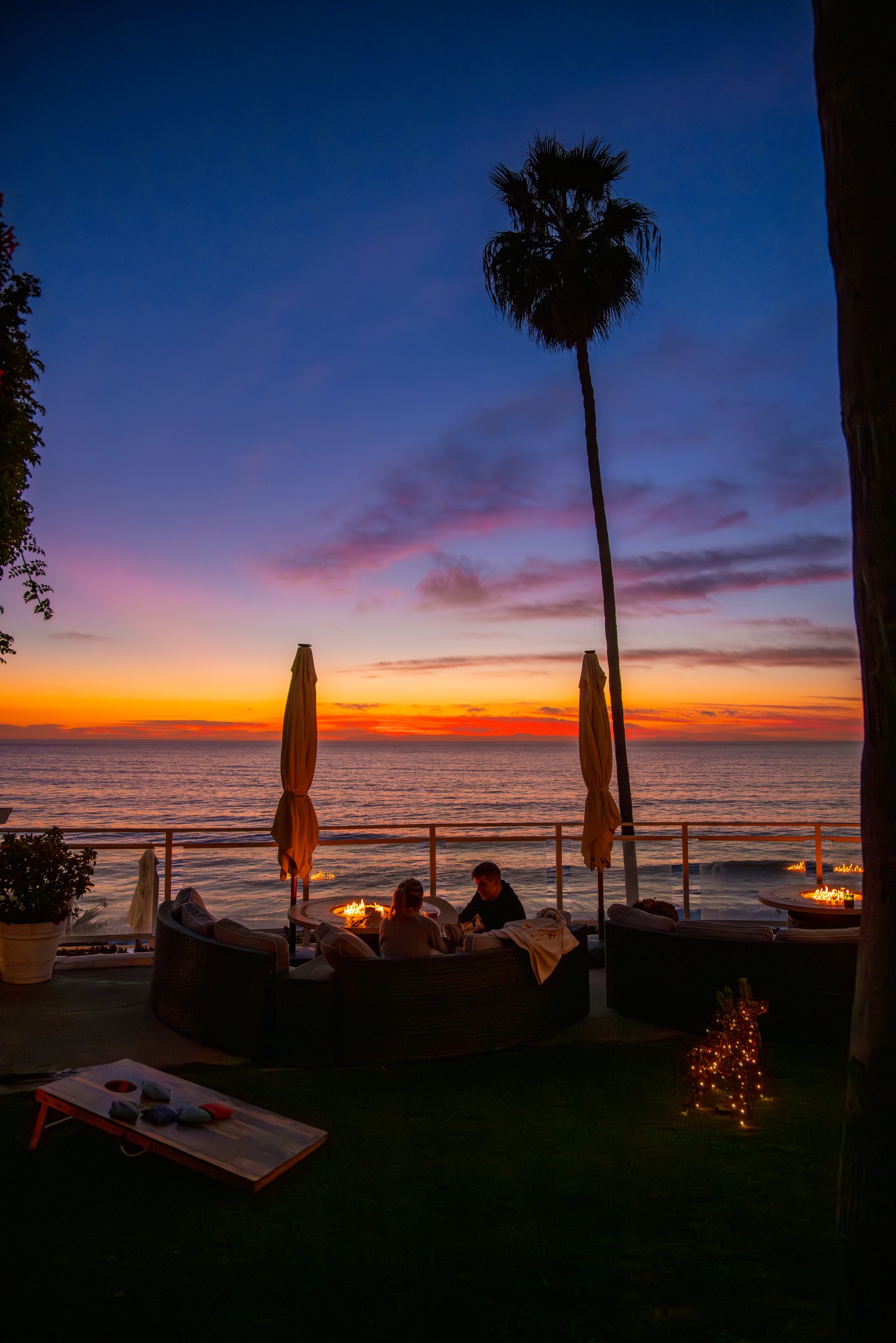 Guests by a fire pit on the lawn at Laguna Surf Lodge as the sun sets over the ocean, with a tall palm above
