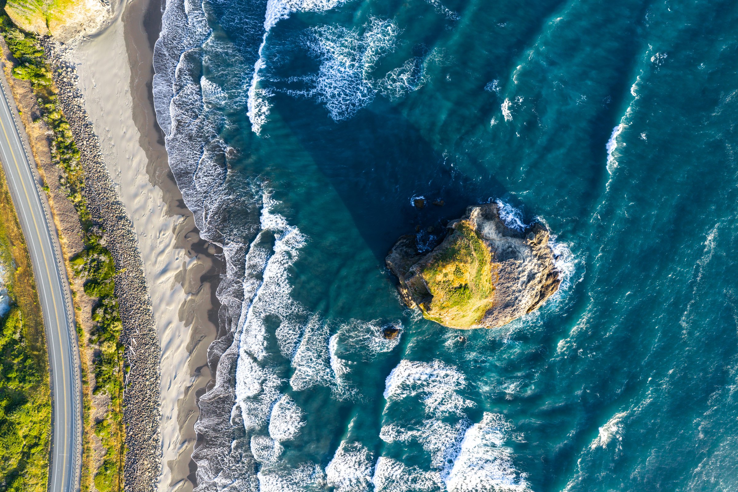 Top down aerial of waves wrapping around a grassy sea stack beside a beach and coastal highway on the central Oregon coast