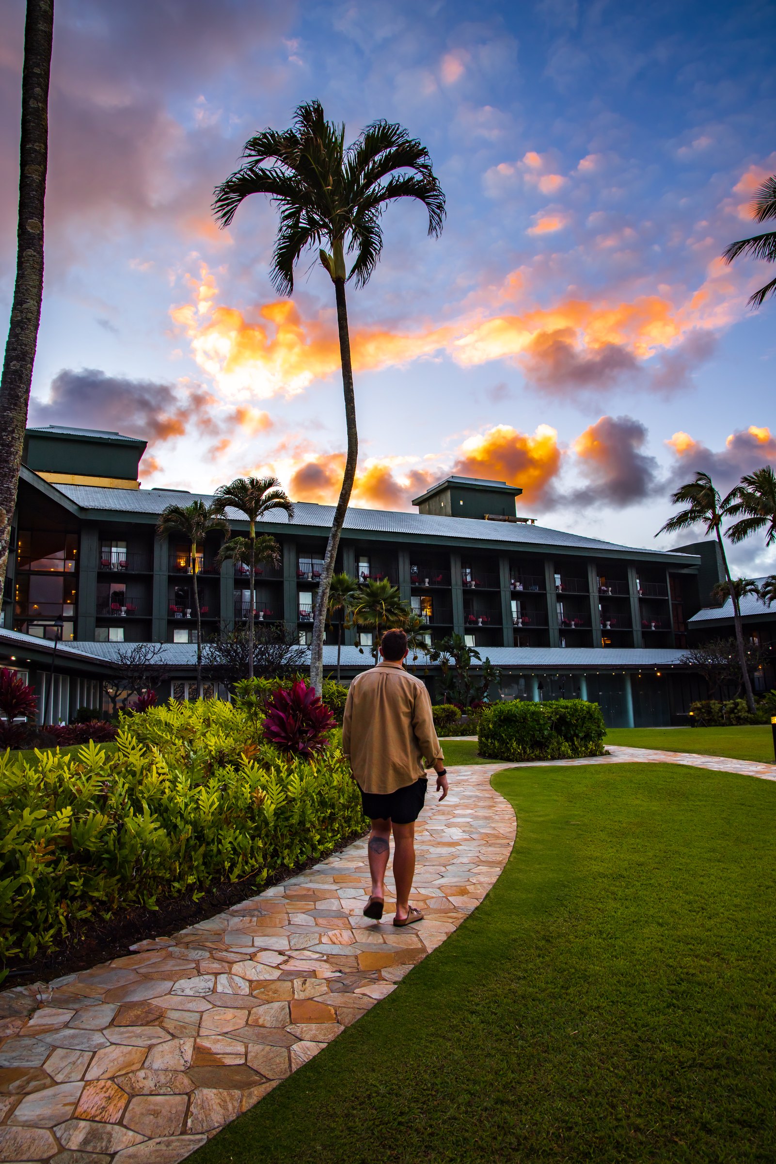 Walking a stone garden path under palms toward Outrigger Kauai Beach Resort at sunset