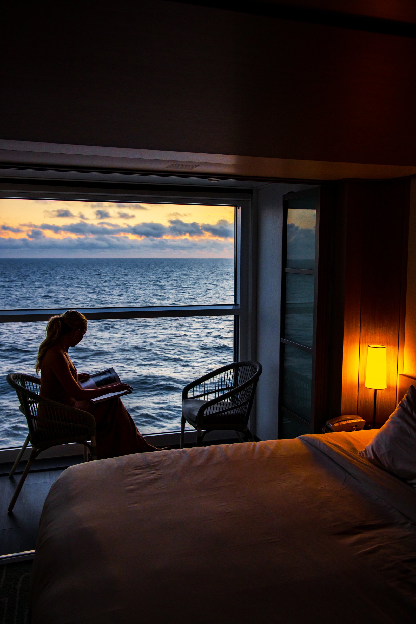 Woman relaxing by a stateroom window looking out at the open ocean at sunset